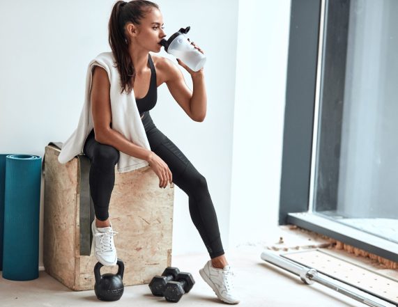 Young woman in leggins with towel on shoulders drinking water after fitness training